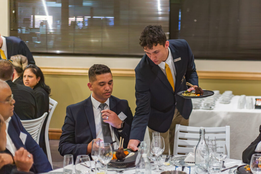A front-of-house hospitality student serving an elegant lamb entree to seated guests at a formal corporate dinner.