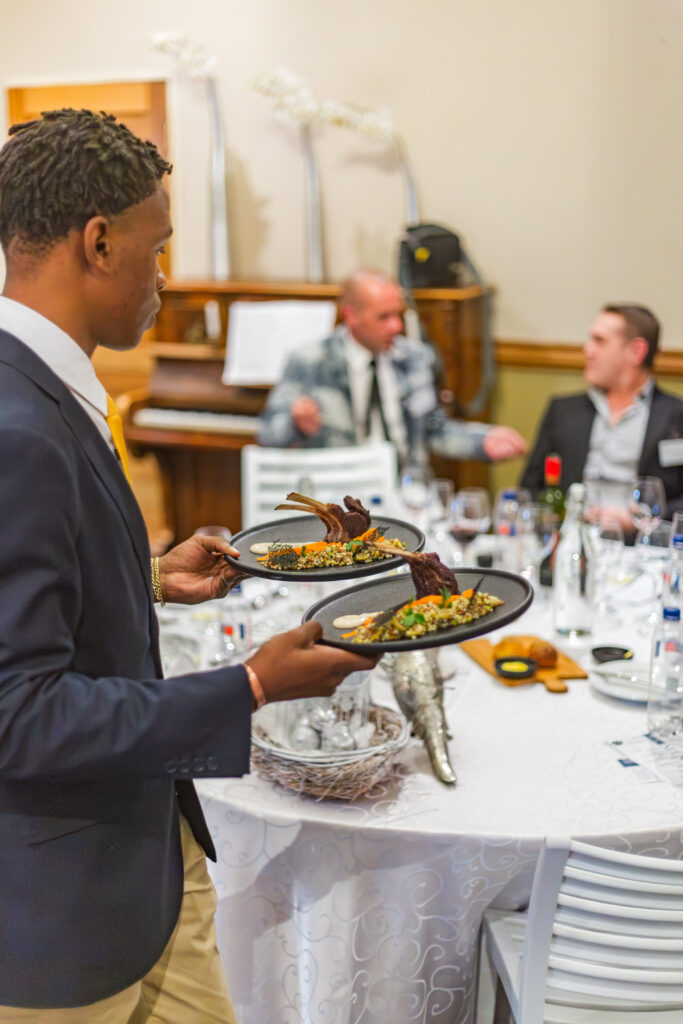 A CHS student carefully serving gourmet lamb entrees to invited industry professionals at a round dining table.