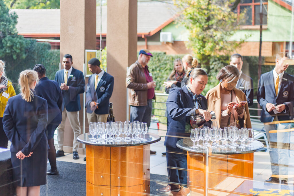 Hospitality students pouring wine and preparing cocktail glasses for guests at an outdoor networking reception table.