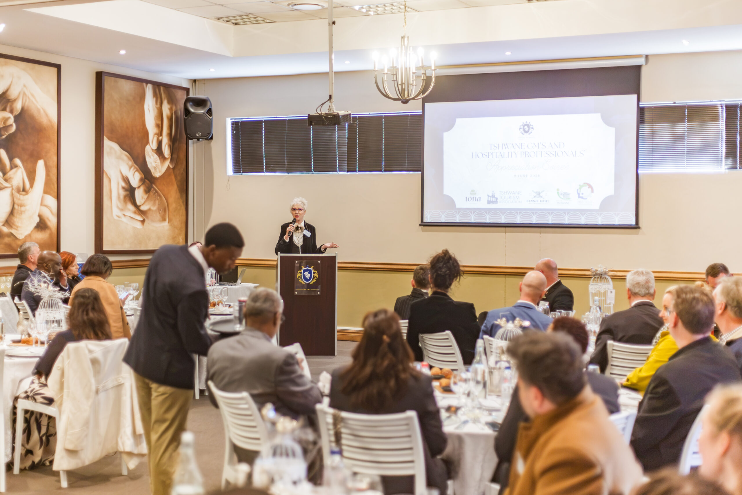 A keynote speaker addressing an audience of hotel general managers and hospitality professionals from a podium in a banquet hall.